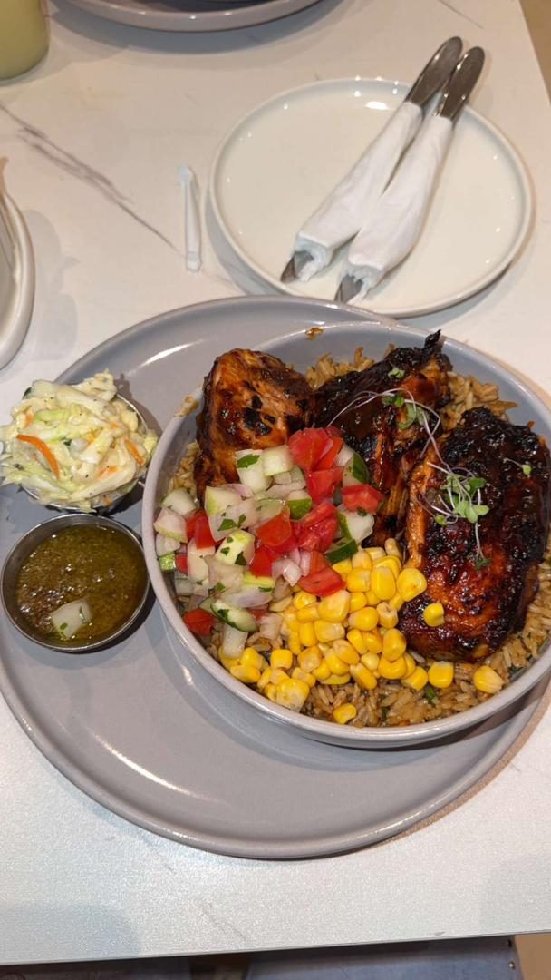A generous restaurant bowl of jollof rice topped with grilled chicken, fresh salsa, corn and coleslaw